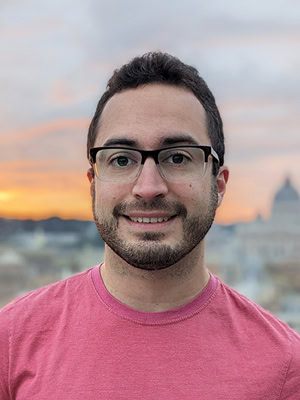 Portrait of Emerson Nolasco against a city sunset in the background. Emerson has dark hair and glasses and is smiling.