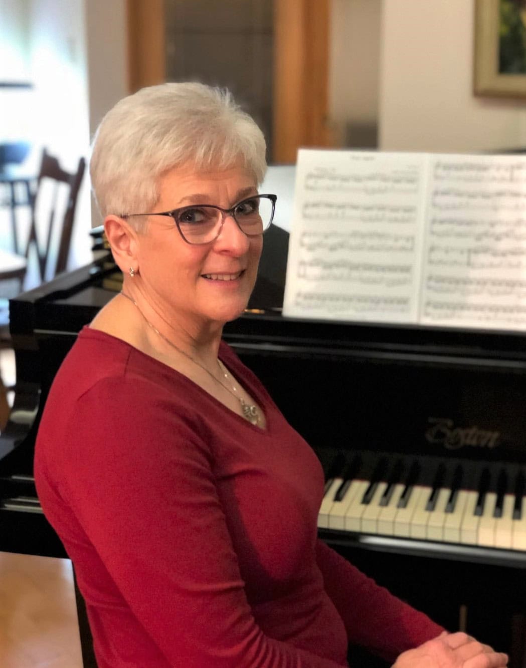 Elaine Krul seated at a piano, turned to face the camera behind her. Elaine has silver hair, dark glasses and is smiling confidently.