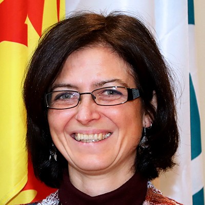 Portrait of Giorgia Purcaro, a woman with short dark hair and glasses, smiling while standing in front of colorful flags.