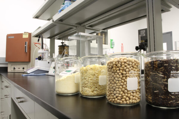 three lab beakers filled with seeds and grains sit on a black counter top with lab shelving and instruments in the background