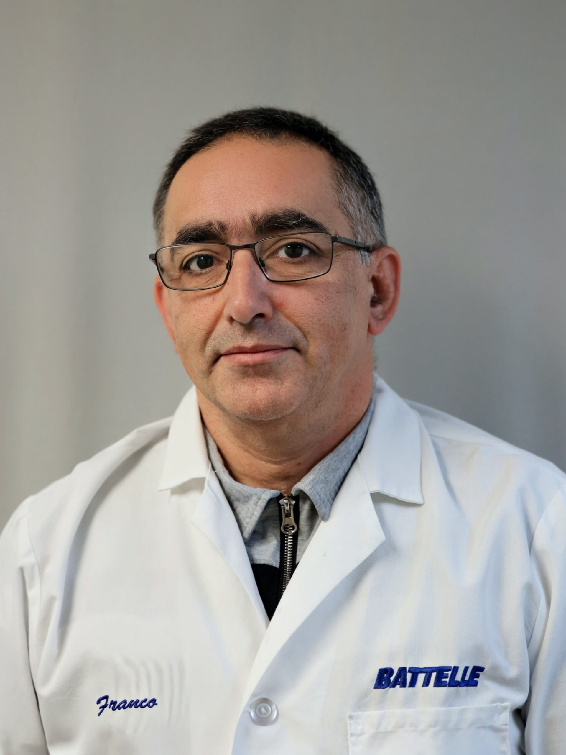 Professional headshot of a person wearing glasses and a white Battelle laboratory coat, seated against a light gray background.