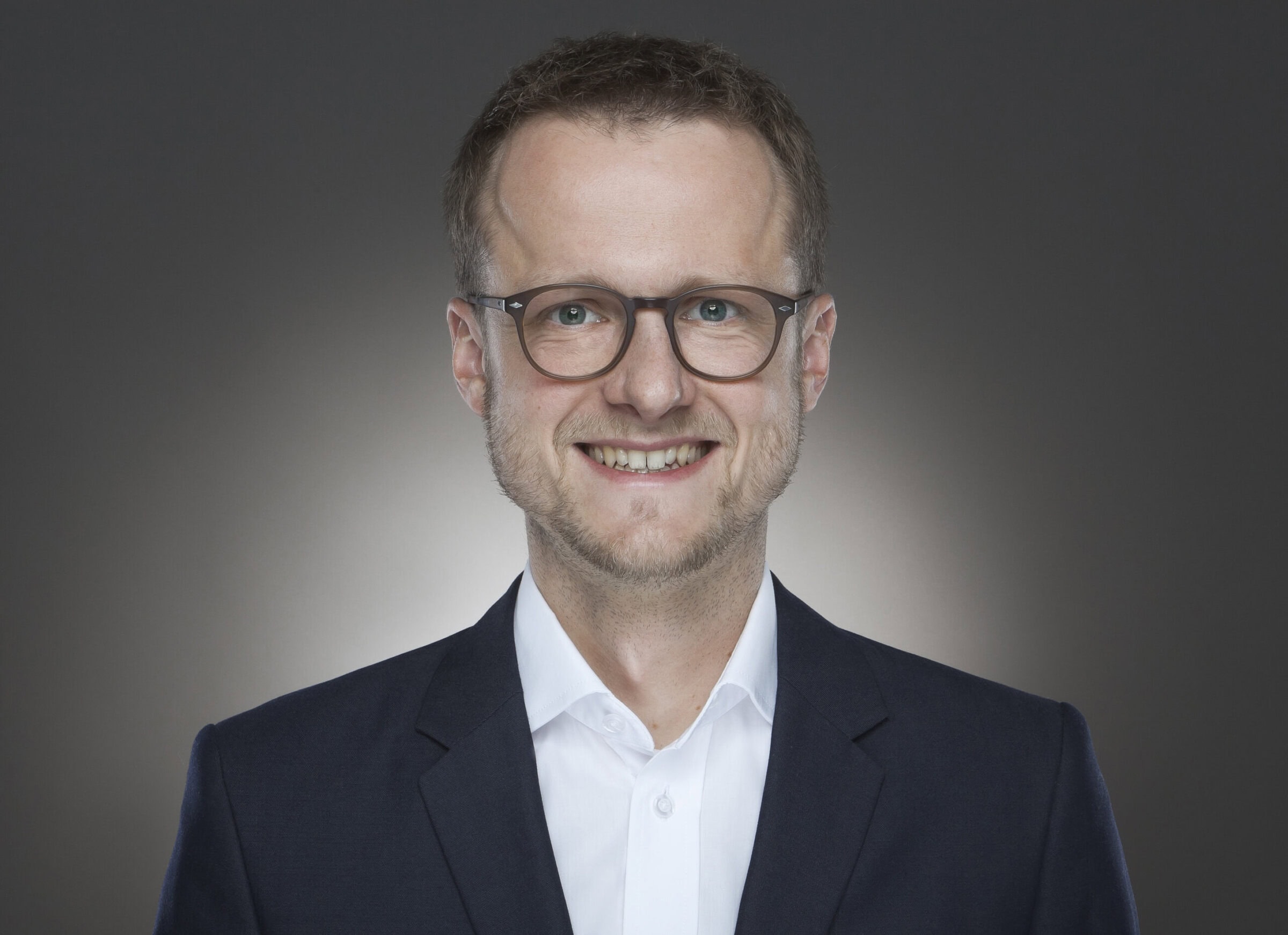 Professional headshot of Jochen Ziegler wearing glasses, a dark blazer, and white shirt, smiling against a neutral gray background.