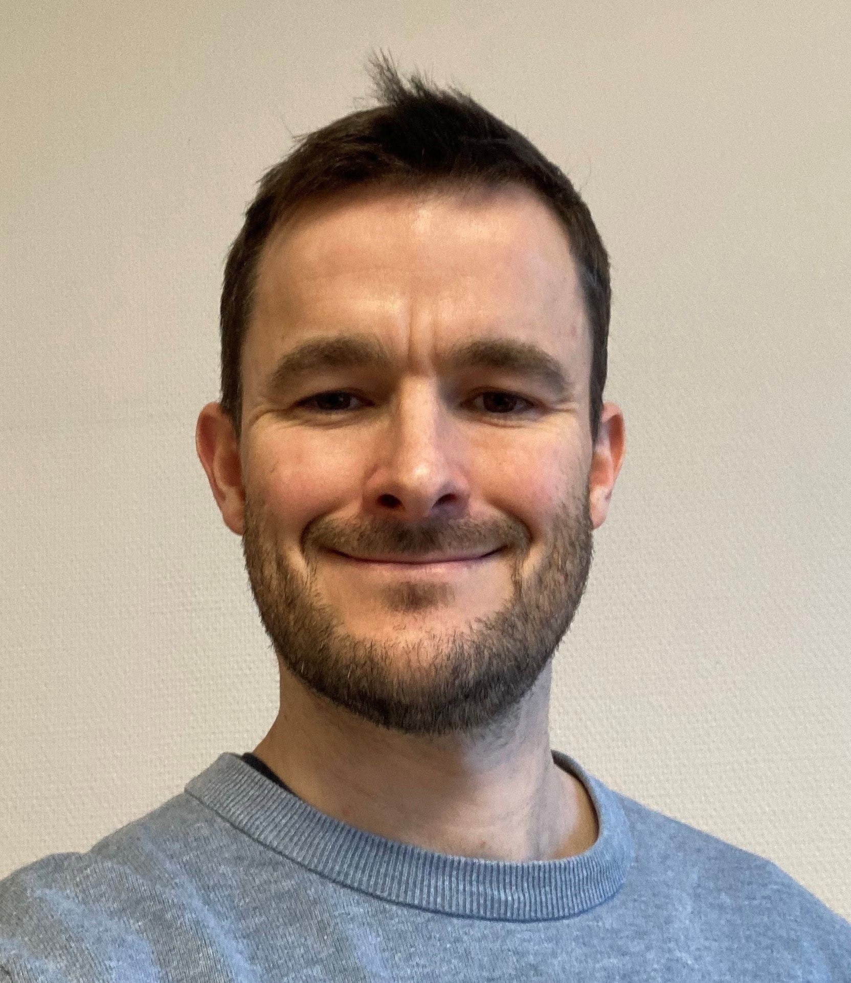 Head-and-shoulders portrait of Ross Ellis wearing a light blue sweater against a neutral background.