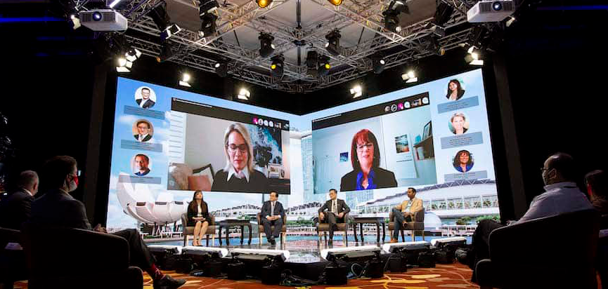 Four speakers on stage at a hybrid event with users on a livestream on a screen behind the stage
